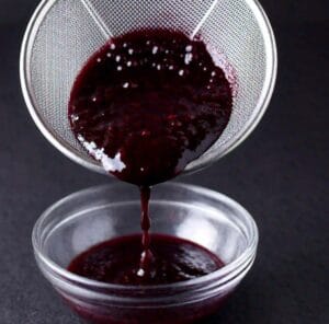 How to Make Blackberry Sauce for Cheesecake? 3 Strained blackberry sauce being poured into a glass bowl, with a sieve catching the seeds.